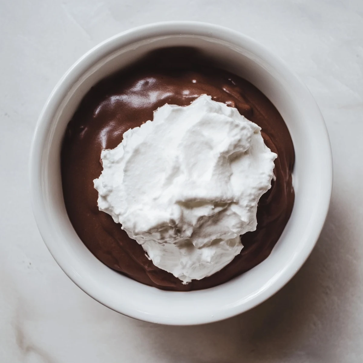 Smooth, glossy chocolate pudding sits in a small glass dish, crowned with a fluffy dollop of homemade whipped cream and shaved chocolate.