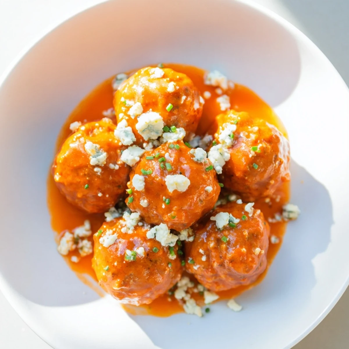 Tender, spicy chicken meatballs glistening with buttery buffalo sauce and garnished with fresh chives. Piled high on a white plate, perfect for a party snack or game day spread. 