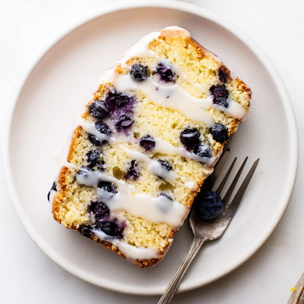 Golden slice of lemon blueberry cake drizzled with white glaze on a white plate